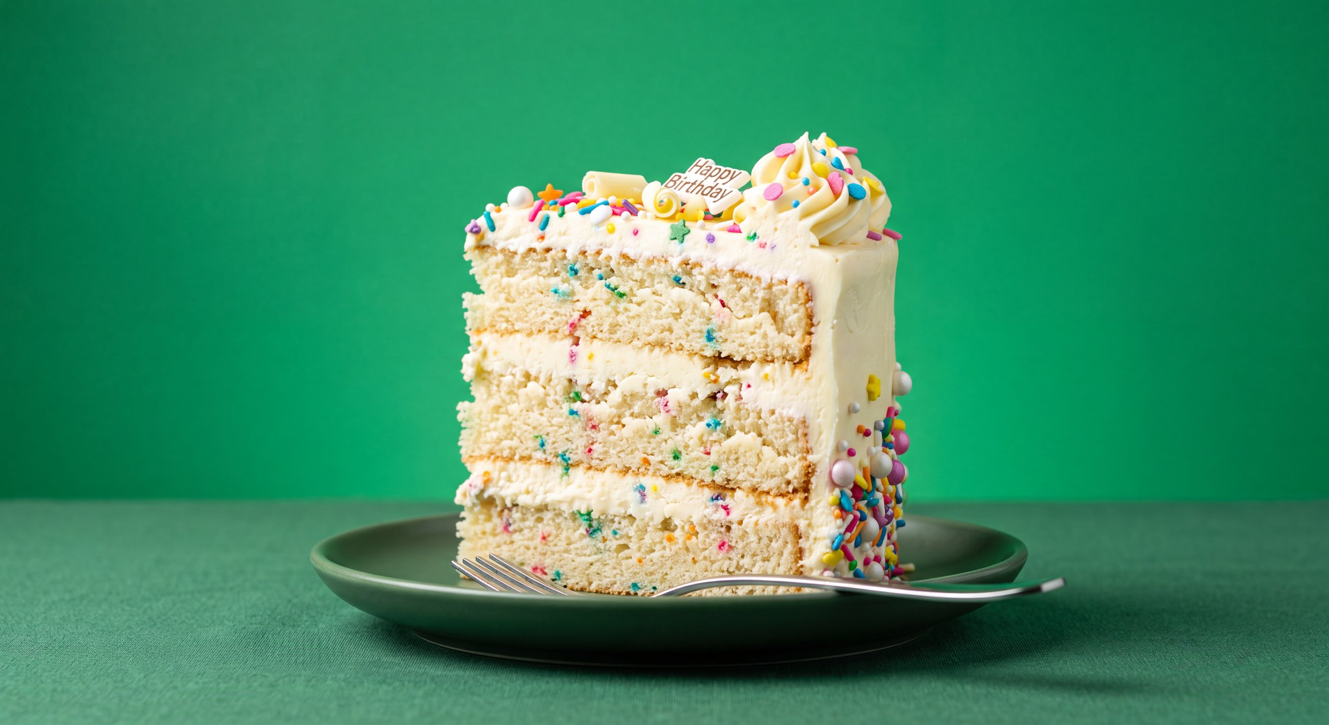 A tall slice of confetti birthday cake with white frosting, sprinkles, and a Happy Birthday plaque, on a green backdrop